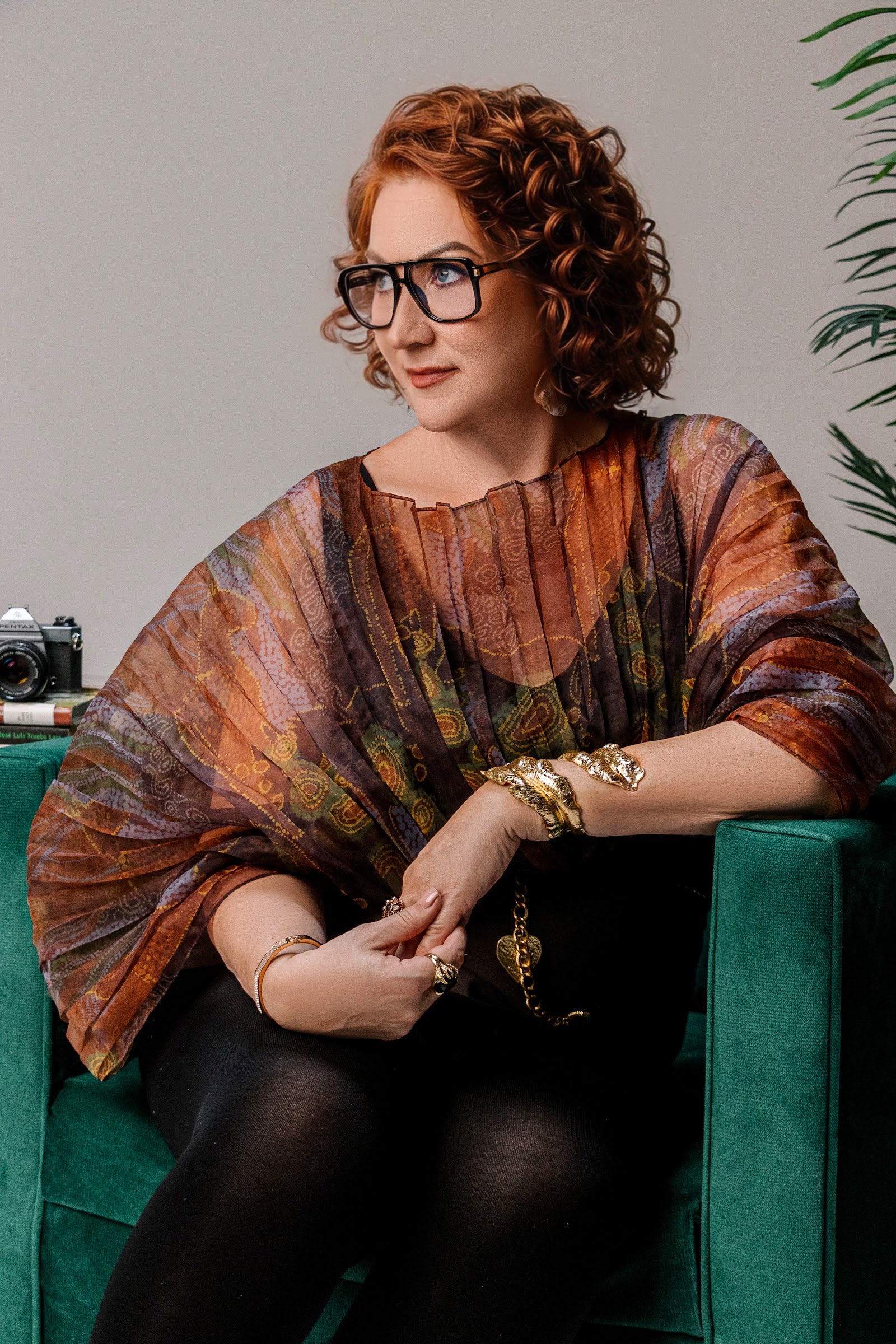 Kelly Diels in a patterned top and glasses, seated in a green velvet chair